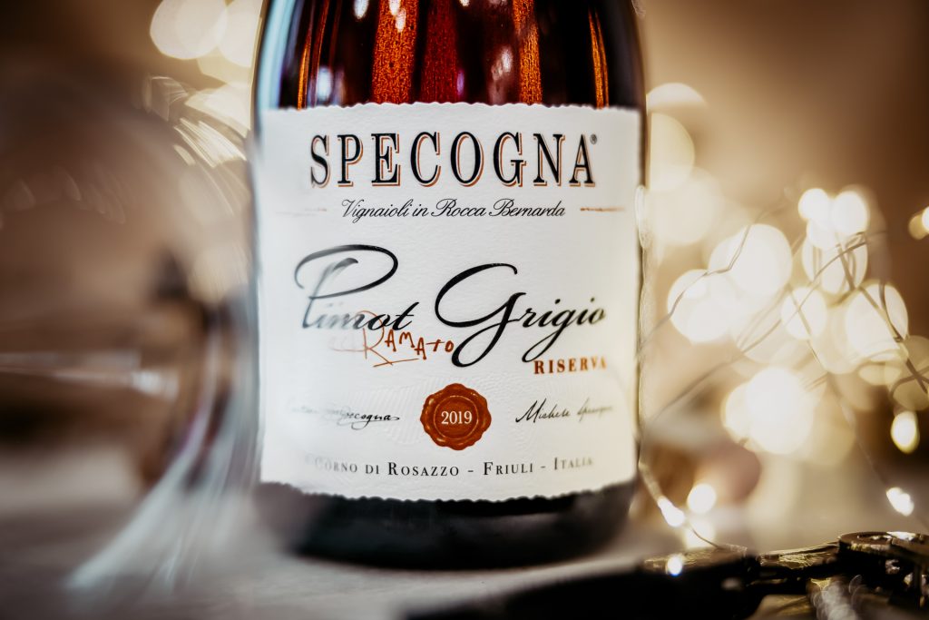 Fotografia detailu bielej etikety vína Specogna Pinot grigio, v popredí rozmazaný pohár na víno, v pozadí rozmazané svetielka